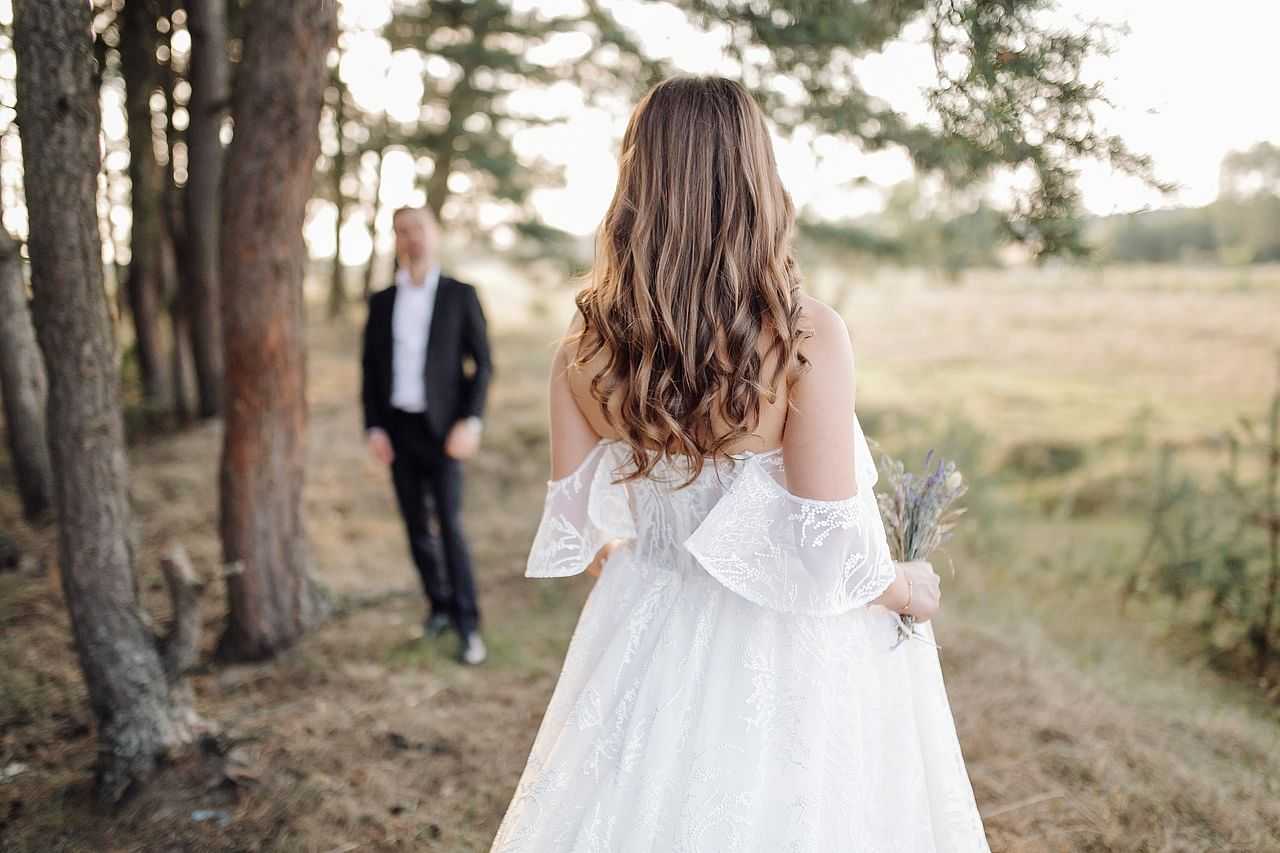 Bride holding flowers facing groom in a sunlit forest clearing.